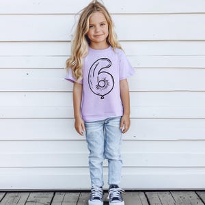 May include: A young person wearing a lavender t-shirt with a black outline of a number six balloon. The shirt is a birthday shirt for a six-year-old.