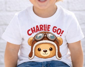 Puede incluir: Camiseta blanca con un oso de peluche de dibujos animados que lleva un gorro de aviador marrón y gafas. El texto rojo "CHARLIE GO!" está impreso encima del oso. La camiseta la lleva un niño.