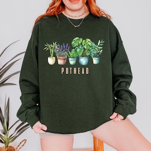 May include: Dark green sweatshirt featuring a watercolor illustration of various potted plants. The word "POTHEAD" is printed in a light brown color below the plants. The sweatshirt is a solid color and has a crew neck.