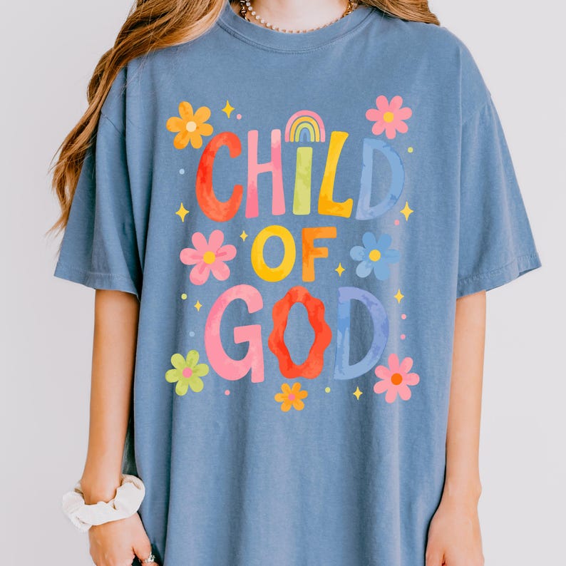 May include: A blue t-shirt with the words "CHILD OF GOD" in colorful, floral-style lettering. The design includes a rainbow and various flower illustrations. The shirt is a casual, short-sleeved style.