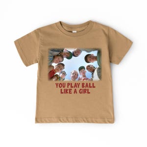 Peut inclure: T-shirt &agrave; manches courtes de couleur beige avec un motif repr&eacute;sentant un groupe de gar&ccedil;ons regardant vers le bas. Le texte "YOU PLAY BALL LIKE A GIRL" est imprim&eacute; sous le motif.