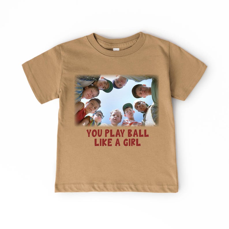 Peut inclure: T-shirt &agrave; manches courtes de couleur beige avec un motif repr&eacute;sentant un groupe de gar&ccedil;ons regardant vers le bas. Le texte "YOU PLAY BALL LIKE A GIRL" est imprim&eacute; sous le motif.