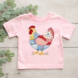 May include: A light pink t-shirt featuring a colorful patchwork chicken design. The chicken has a red comb, yellow beak, and a body composed of various patterned fabric pieces, including gingham, floral, and butterfly motifs. The shirt is made of soft material.
