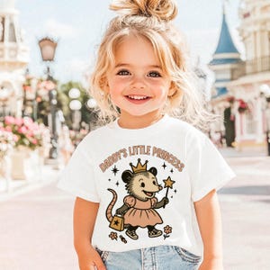 May include: A white short-sleeve t-shirt featuring a graphic of an opossum wearing a crown and a dress, holding a star wand and a bag. The text "DADDY'S LITTLE PRINCESS" is printed in an arch above the opossum, accompanied by small star and flower icons.