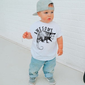 May include: A white t-shirt with a black and white illustration of an opossum and the word "AWESOME" printed on it. A young child is wearing the shirt with blue jeans and a gray baseball cap.