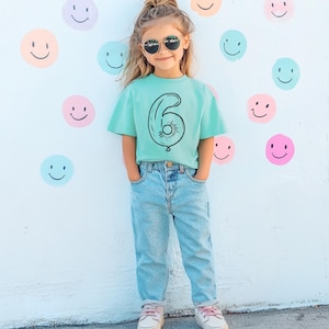 May include: A child wearing a turquoise t-shirt with a black outline of a number six balloon. The child is wearing blue jeans and sunglasses.