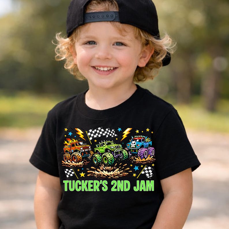 Może przedstawiać: Czarny t-shirt z trzema kolorowymi monster truckami, z szachownicami i błyskawicami. Tekst "TUCKER'S 2ND JAM" jest wydrukowany na zielono. Koszulka jest noszona przez uśmiechnięte dziecko.