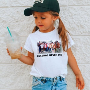 May include: A white t-shirt with a graphic of a group of boys playing baseball. The graphic is surrounded by the text "LEGENDS NEVER DIE".