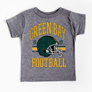 Puede incluir: Camiseta gris con un dise&ntilde;o de casco de f&uacute;tbol y las palabras "GREEN BAY FOOTBALL" en amarillo. El casco es verde y blanco, con el n&uacute;mero "19" a cada lado.