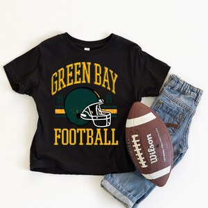 Puede incluir: Camiseta negra con el texto "GREEN BAY FOOTBALL" y un gr&aacute;fico de casco verde. Un bal&oacute;n de f&uacute;tbol marr&oacute;n y blanco y unos vaqueros azules est&aacute;n al lado de la camiseta. La camiseta tiene un dise&ntilde;o de estilo vintage.