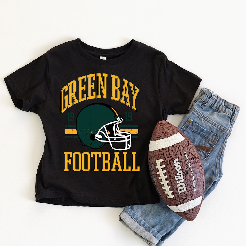 Puede incluir: Camiseta negra con el texto "GREEN BAY FOOTBALL" y un gr&aacute;fico de casco verde. Un bal&oacute;n de f&uacute;tbol marr&oacute;n y blanco y unos vaqueros azules est&aacute;n al lado de la camiseta. La camiseta tiene un dise&ntilde;o de estilo vintage.