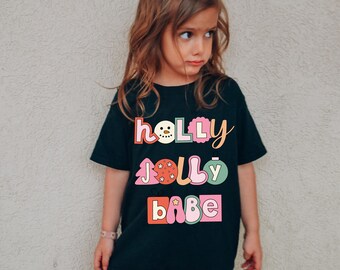 May include: Black t-shirt with the words "HOLLY JOLLY BABE" in colorful, retro-style lettering. The design includes a snowman, Christmas tree, ornament, and star accents. The shirt is worn by a young child.