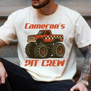 Pu&ograve; includere: T-shirt color crema con grafica di monster truck rosso e la scritta "Cameron's Pit Crew" in arancione. Il camion ha pneumatici grandi e neri e un motivo a scacchi sul cassone.