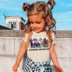 May include: A young girl wearing a light yellow t-shirt with a vintage photo of a group of boys playing baseball. The shirt has the text "Legends Never Die" printed on the front. The girl is wearing a black and white plaid skirt and has her hair pulled back in two pigtails.