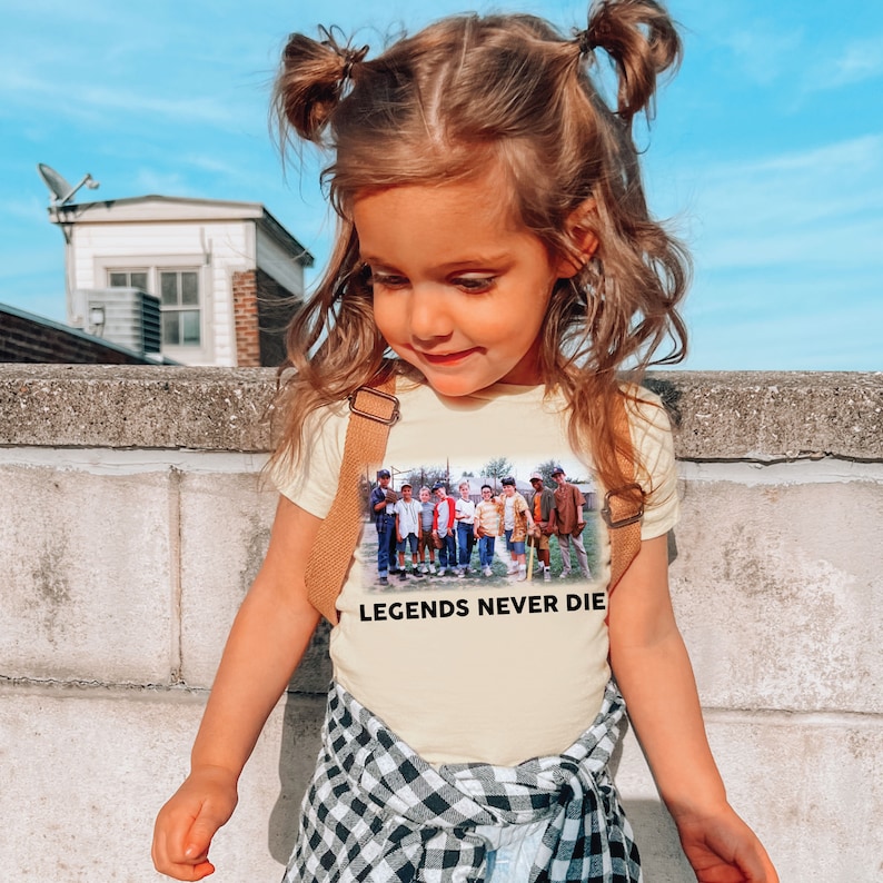 May include: A young girl wearing a light yellow t-shirt with a vintage photo of a group of boys playing baseball. The shirt has the text "Legends Never Die" printed on the front. The girl is wearing a black and white plaid skirt and has her hair pulled back in two pigtails.