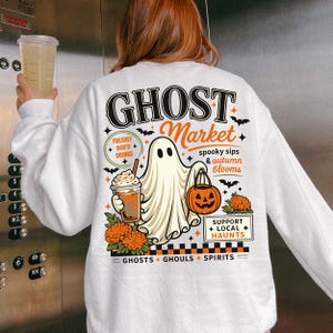 May include: A white long-sleeve sweatshirt featuring a back graphic of a ghost holding a drink and a pumpkin bucket. The text reads GHOST Market, FRESHLY BOO'D DRINKS, spooky sips & autumn blooms, SUPPORT LOCAL HAUNTS, and GHOSTS GHOULS SPIRITS.