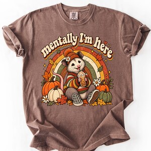 May include: A brown short-sleeve t-shirt featuring a graphic of an opossum wearing a beanie and sweater, holding a drink. The text reads 'mentally I'm here' above a rainbow, pumpkins, and autumn leaves. The shirt has a crew neck and rolled sleeves.