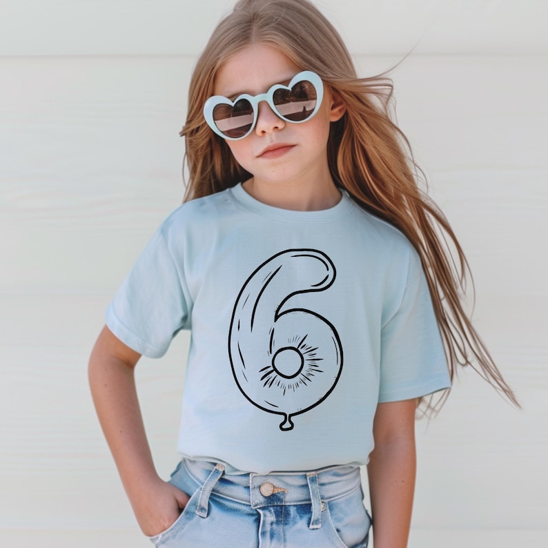May include: A light blue t-shirt with a black outline of a number six shaped like a balloon. The shirt is being worn by a young girl with long blonde hair wearing heart-shaped sunglasses.