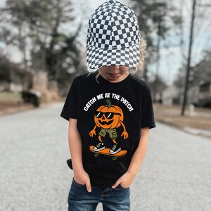 May include: A black t-shirt featuring a graphic of an orange pumpkin character wearing sunglasses, green shorts, and black sneakers while riding a skateboard. The text CATCH ME AT THE PATCH is printed above the pumpkin. A black and white checkered baseball cap.