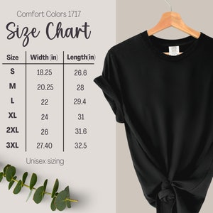 May include: A black t-shirt hanging on a wooden hanger, displayed next to a Comfort Colors 1717 size chart. The chart lists sizes S through 3XL with corresponding width and length measurements in inches. The shirt features a knotted hem at the bottom.