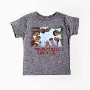 Peut inclure: Un t-shirt gris chin&eacute; avec un motif repr&eacute;sentant un groupe d'enfants regardant la cam&eacute;ra. Le texte "You play ball like a girl" est imprim&eacute; sous l'image.