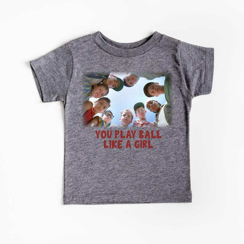 Peut inclure: Un t-shirt gris chin&eacute; avec un motif repr&eacute;sentant un groupe d'enfants regardant la cam&eacute;ra. Le texte "You play ball like a girl" est imprim&eacute; sous l'image.