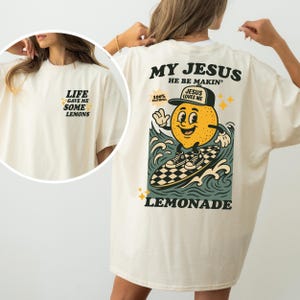 May include: Cream-colored t-shirt with the text "LIFE GAVE ME SOME LEMONS" on the front and a cartoon lemon surfing on the back. The back graphic includes the text "MY JESUS HE BE MAKIN' LEMONADE".