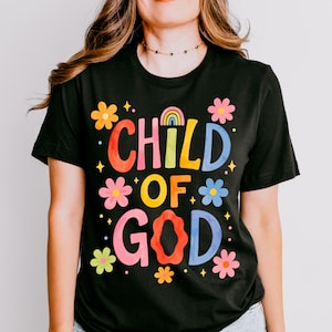 May include: Black t-shirt with the words "CHILD OF GOD" in colorful, bubble letters. The design includes flowers and a rainbow. The shirt is short-sleeved.