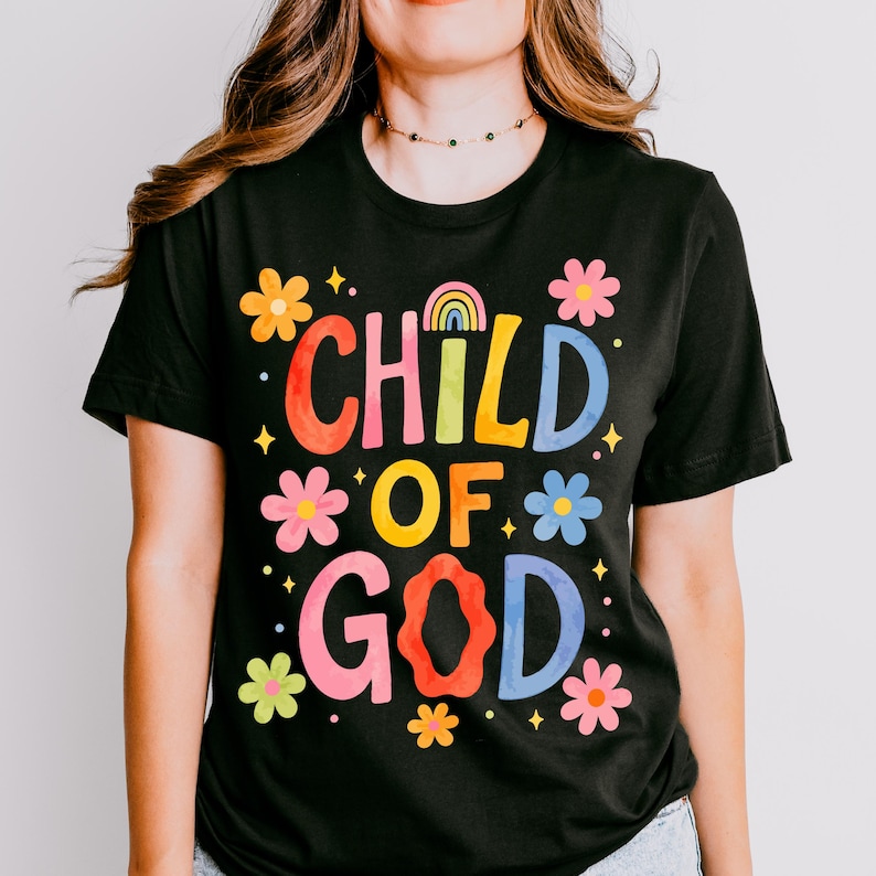May include: Black t-shirt with the words "CHILD OF GOD" in colorful, bubble letters. The design includes flowers and a rainbow. The shirt is short-sleeved.
