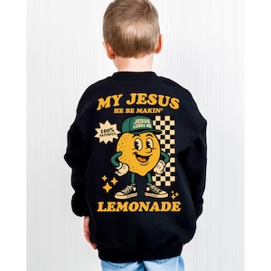 May include: Black sweatshirt with a cartoon lemon character wearing a baseball cap that says "Jesus Loves Me." The sweatshirt has the text "My Jesus He Be Makin' Lemonade" in yellow and white. The design includes stars and a checkered flag.