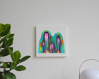May include: A framed abstract painting with a blue, green, yellow, and pink color scheme. The painting is hanging on a white wall above a wooden table.