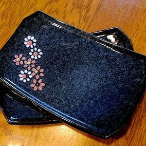 May include: Two dark blue ceramic rectangular plates with a floral design. The plates have a speckled glaze and are decorated with white and pink flowers.