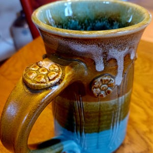 May include: A handmade ceramic mug with a unique design. The mug features a brown handle with decorative floral accents and a blue, brown, and green color scheme. The interior is a deep green, and the mug has a textured, glazed finish.