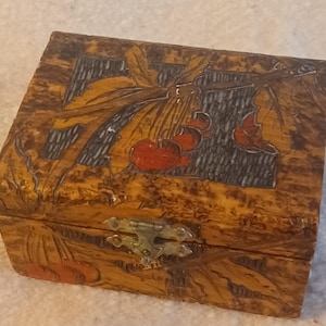 May include: A rectangular wooden box with a hinged lid featuring a carved and painted design of red cherries on a branch against a dark textured background. The box has a light brown mottled finish and a silver-toned metal latch on the front.