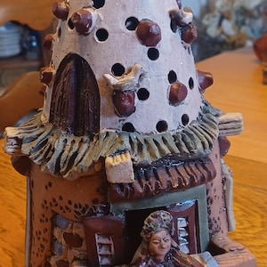 May include: A detailed ceramic figurine of a small house or tower. The structure is primarily tan and brown, with a conical top dotted with dark brown protrusions. The base features a seated figure in a window. The roof has a thatched appearance.