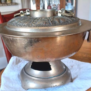 May include: A vintage brass cooking pot with a decorative lid featuring embossed patterns. The pot has a wide, shallow bowl shape and sits on a pedestal base. The lid has ornate handles. The pot is resting on a white cloth.