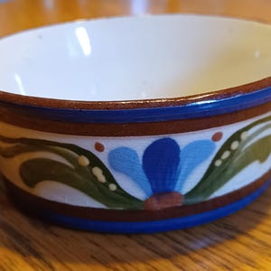 May include: A small ceramic bowl with two side handles, featuring a white interior and a cream-colored exterior band decorated with hand-painted blue and green floral motifs, framed by dark brown and blue horizontal stripes along the rim and base.