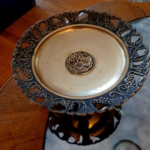 May include: An antique brass-toned serving tray with a decorative grape design. The tray has a central medallion with a grape cluster and leaf, surrounded by an oval surface. The outer rim features a detailed grape vine pattern.