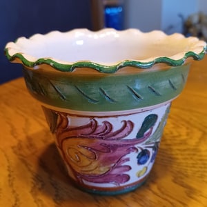 May include: A ceramic planter featuring a scalloped rim with a green painted edge. The upper section has a green band with carved diagonal lines. The body is decorated with hand-painted floral motifs in shades of pink, yellow, and blue on white.