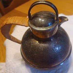 May include: A silver-toned, vintage tea or coffee pot with a woven handle. The pot features an ornate floral pattern and a lid with a small handle. The handle is wrapped in a light brown woven material.