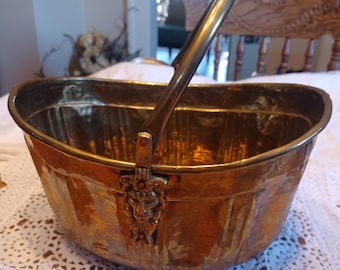 May include: A vintage brass bucket with a handle. The bucket has a hammered texture and a decorative lion's head detail on the handle attachment. The handle is arched and made of brass. The bucket is oval-shaped.