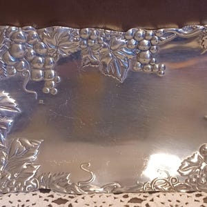 May include: Silver-colored rectangular serving tray with embossed grape and leaf design around the edges. The tray has a smooth, reflective surface in the center. The corners feature detailed grape clusters and foliage.