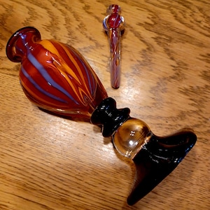 May include: A set of three glass pieces. The largest piece is a red, orange, and blue striped vase. The second piece is a clear glass with red and purple stripes. The third piece is a black and clear glass piece.