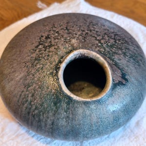 May include: A round, ceramic vase with a dark, mottled glaze in shades of gray, green, and brown. The vase has a wide, open mouth and a textured surface. The overall shape is flat and disc-like.