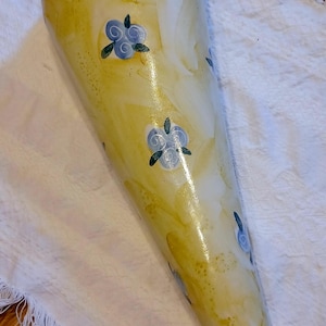 May include: A yellow ceramic wall vase with a cone shape and blue floral pattern. The vase is sitting on a white fringed fabric.