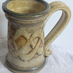 May include: A handcrafted stoneware mug with a large handle. The mug features a textured, woven design on the body and a dark rim. The color palette includes shades of tan, brown, and cream, giving it an earthy aesthetic.