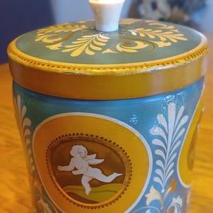 May include: A vintage blue and yellow tin container with a white knob on the lid. The container features a decorative design with a cherub in a gold frame. The lid has a gold and yellow floral pattern.