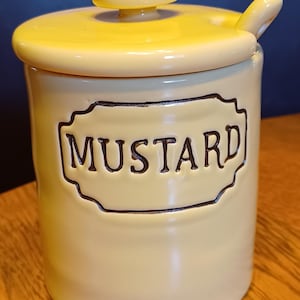 May include: A pale yellow ceramic mustard jar with a matching lid and a small spoon handle protruding from the side. The front features a decorative embossed label with a dark brown outline containing the word MUSTARD in dark brown lettering.