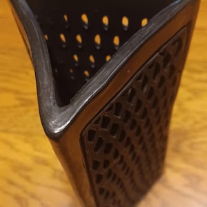 May include: A tall, triangular black vase featuring a lattice-style cutout pattern on its sides. The vase has a smooth, dark rim and a hollow interior, with the intricate geometric openwork design extending down the length of the exterior walls.