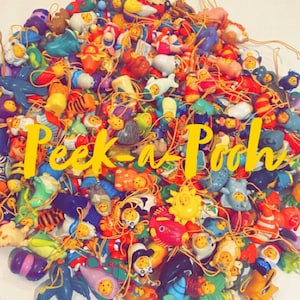 Può includere: Una grande collezione di ciondoli di plastica colorati con i personaggi di Winnie the Pooh. I ciondoli sono attaccati a delle stringhe e hanno una varietà di disegni, tra cui Winnie the Pooh, Piglet, Tigger, Eeyore e Owl. I ciondoli sono disposti in un mucchio e il testo "Peek-a-Pooh" è visibile al centro dell'immagine.